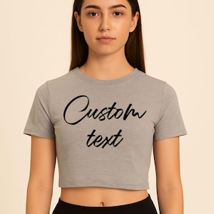 May include: A grey crop top with short sleeves and the words "Custom text" in black script. The shirt is a light grey colour and has a round neckline. The Custom247 logo is in the top left corner.