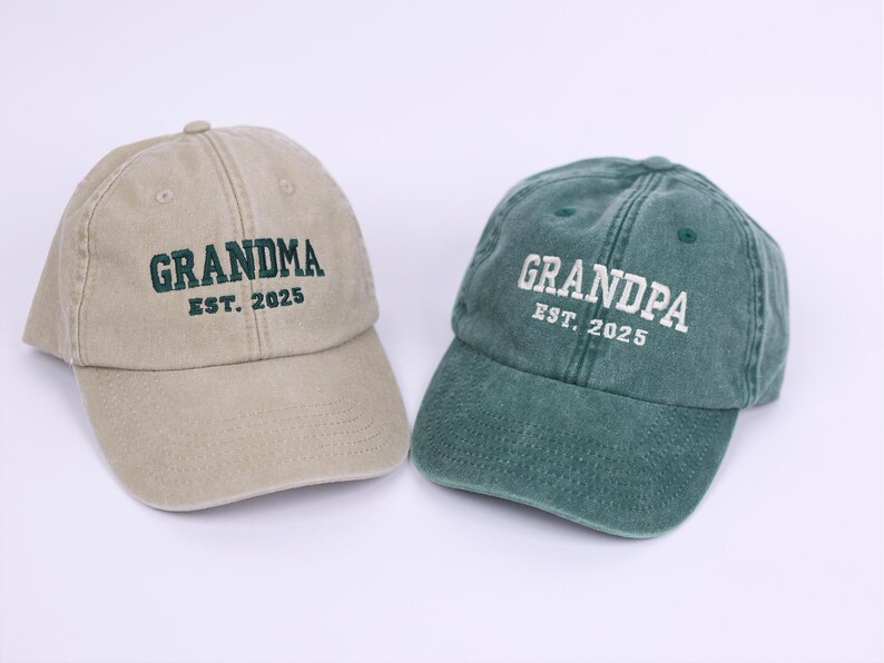 May include: Two baseball caps, one tan and one green, with embroidered text. The tan cap says "Grandma Est. 2025" and the green cap says "Grandpa Est. 2025".