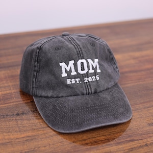May include: A black denim baseball cap with white embroidered text that reads "MOM EST. 2025".