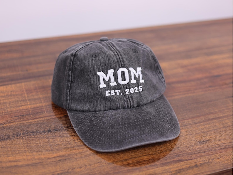 May include: A black denim baseball cap with white embroidered text that reads "MOM EST. 2025".