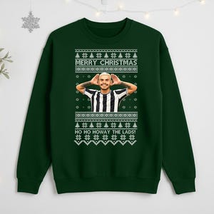 May include: A dark green Christmas sweater with a festive design. The sweater features the text "MERRY CHRISTMAS" and "HO HO HOWAY THE LADS!" in white. A person is in the center of the sweater. The sweater has a crew neck and long sleeves.