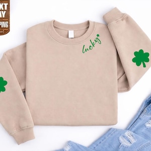 Puede incluir: Sudadera beige con trébol verde y texto "Lucky". La sudadera tiene cuello redondo y mangas largas. Un trébol verde está en cada manga. Vaqueros azules en el fondo.