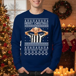 May include: Navy blue Christmas sweater with a festive design. The sweater features the text "MERRY CHRISTMAS" and "HO HO HOWAY THE LADS!" along with a graphic of a person. The design includes snowflakes and Christmas trees.