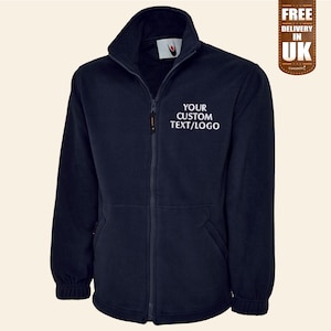 Könnte beinhalten: Eine marineblaue Fleecejacke mit Reißverschluss und zwei Taschen. Die Jacke hat ein weiß gesticktes Design mit der Aufschrift "YOUR CUSTOM TEXT/LOGO".