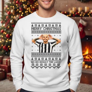 May include: White Christmas sweater with a festive design. The sweater features a graphic of a person with their hands up, and the text "MERRY CHRISTMAS" and "HO HO HOWAY THE LADS!" are displayed across the front. The sweater has a crew neck and long sleeves.