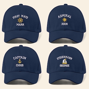 May include: Four navy blue baseball caps with white text and nautical-themed graphics. Each cap features a different title: "First Mate," "Admiral," "Captain," and "Steuermann," along with a name. The caps have a curved brim.