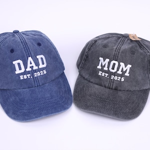May include: Two denim baseball caps, one blue and one gray, with white embroidered text. The blue cap says "DAD EST. 2025" and the gray cap says "MOM EST. 2025".