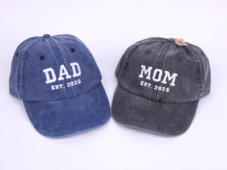 May include: Two denim baseball caps, one blue and one gray, with white embroidered text. The blue cap says "DAD EST. 2025" and the gray cap says "MOM EST. 2025".