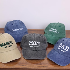 May include: Five different colored baseball caps with embroidered text. The caps are beige, dark gray, green, blue, and denim blue. The text on the caps reads "Grandma Est. 2025", "Mom Est. 2025", "Custom Embroidery", "Dad Est. 2025", and "Custom Text".