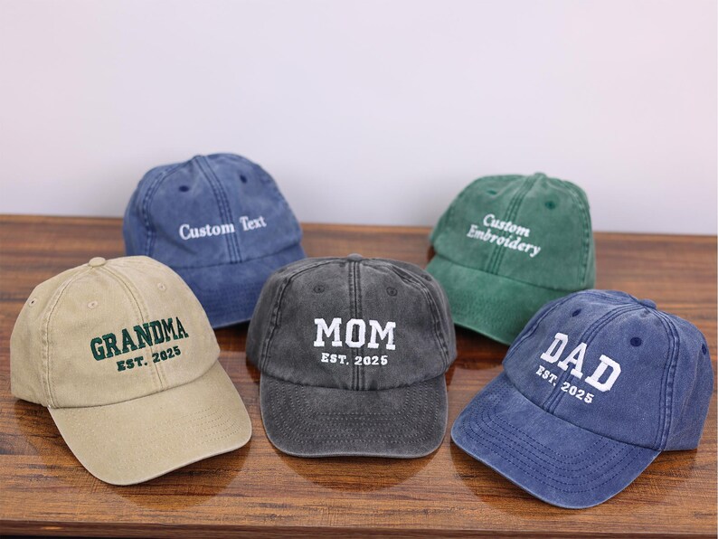 May include: Five different colored baseball caps with embroidered text. The caps are beige, dark gray, green, blue, and denim blue. The text on the caps reads "Grandma Est. 2025", "Mom Est. 2025", "Custom Embroidery", "Dad Est. 2025", and "Custom Text".