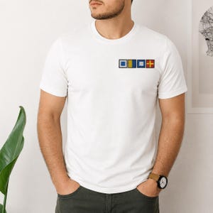 Könnte beinhalten: Ein weißes T-Shirt mit kurzen Ärmeln und einer kleinen Grafik auf der linken Brust, die aus vier quadratischen nautischen Signalflaggen besteht. Die Flaggen sind horizontal angeordnet und zeigen Blau, Gelb, Weiß sowie ein rotes und gelbes Kreuzmuster.