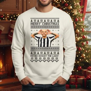 May include: A white Christmas sweater with a festive design. The sweater features the text "MERRY CHRISTMAS" and "HO HO HOWAY THE LADS!". It has a graphic of a person in a black and white striped shirt.