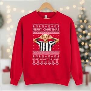 May include: A red Christmas sweatshirt with a festive design. The sweatshirt features the text "MERRY CHRISTMAS" and "HO HO HOWAY THE LADS!" in white, along with a graphic of a person wearing a black and white striped shirt. The design includes snowflakes and Christmas trees.