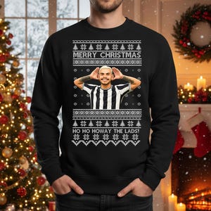 May include: Black Christmas sweater with a festive design. The sweater features the text "MERRY CHRISTMAS" and "HO HO HOWAY THE LADS!" along with a picture of a person. The sweater has a classic crew neck.