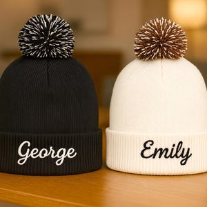 Puede incluir: Dos gorros de punto, uno negro y otro crema, cada uno con un pompón. El gorro negro tiene un pompón blanco y negro y el nombre "George" bordado en blanco. El gorro crema tiene un pompón marrón y el nombre "Emily" bordado en negro.