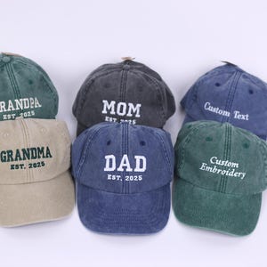 May include: Six different colored baseball caps with embroidered text. The caps are green, blue, grey, tan, and two shades of green. The text on the caps reads "Grandpa Est. 2025", "Mom Est. 2025", "Custom Text", "Grandma Est. 2025", "Dad Est. 2025", and "Custom Embroidery".