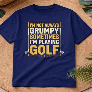 May include: Navy blue t-shirt with the text "I'M NOT ALWAYS GRUMPY SOMETIMES I'M PLAYING GOLF" in a yellow and white design. The shirt features golf club graphics and stars. The tee shirt is displayed on a woven surface.