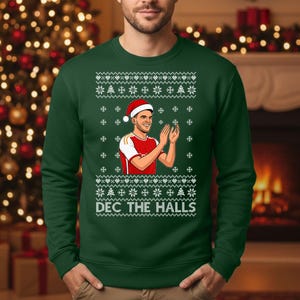 以下が含まれることがあります： お祝いのデザインが施された濃い緑色のクリスマスセーター。セーターには、サンタ帽をかぶって拍手をしている人のグラフィックと、白い文字で「DEC THE HALLS」が描かれています。雪の結晶模様も含まれています。