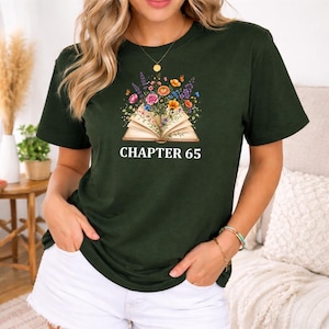 Pode incluir: Uma t-shirt de manga curta verde-escura com um gráfico de um livro aberto com flores coloridas a sair das páginas e o texto "CHAPTER 65" impresso a branco por baixo da ilustração. A t-shirt tem um decote redondo e um corte descontraído.