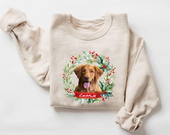 May include: A beige sweatshirt featuring a watercolor illustration of a golden retriever dog within a Christmas wreath. The wreath includes red berries and green leaves. The dog's name, "Charlie", is on a red banner.