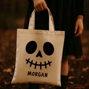 Puede incluir: Una bolsa de lona beige con un diseño de cara de calavera negra. La calavera tiene ojos grandes y redondos, una nariz triangular y una boca cosida. El nombre "MORGAN" está impreso en negro debajo. La bolsa tiene dos asas.