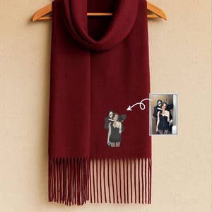 May include: A burgundy scarf with a fringe hem, hanging on a wooden hanger. The scarf has a small graphic of a person and a ghost-faced figure. A small photo of the same figures is attached to the scarf.