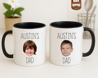 Op de afbeelding: Twee witte keramische mokken met zwarte handvatten en zwarte binnenkant. Elke mok heeft een foto van een kindergezicht, de tekst "AUSTIN'S" boven de foto en het woord "DAD" eronder. De mokken zijn gepersonaliseerd.