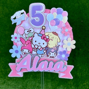May include: Pink and purple cake topper featuring a Hello Kitty character, a frog, a dog, and an aeroplane. The topper also includes the number 5, balloons, and the name "Alaia".