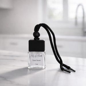 May include: A small, clear glass bottle with a black square cap and a black cord. The bottle has the text "Fitz & Flood" and "Your Scent" printed on it. The cord is attached to the top of the bottle and has a wooden bead.