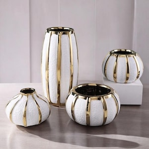 May include: Set of four white ceramic vases with gold accents. The vases are in different sizes and shapes, with a tall cylindrical vase, a round vase, and two smaller round vases.