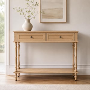 May include: A light brown wooden console table featuring two drawers with small round knobs, turned legs, and a lower shelf. A white ceramic vase containing green leafy branches sits on the top surface of the rectangular wooden table.