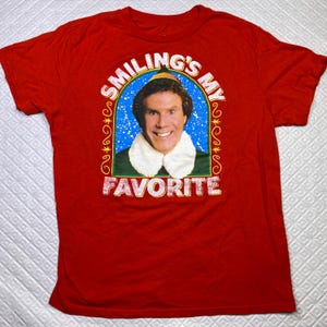 May include: Red t-shirt featuring a smiling image of a man in an elf costume. The text "SMILING'S MY FAVORITE" is printed above and below the image in a white, stylized font. The shirt is made of a soft material.