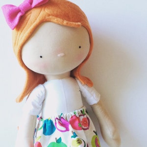 May include: A handmade doll with orange hair, a pink bow, and a white dress with a colourful fruit print.