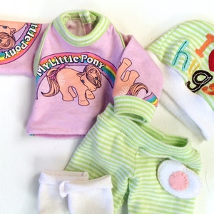 May include: A pink and green baby outfit with a My Little Pony graphic on the shirt. The outfit includes a long-sleeved shirt, trousers, socks, and a hat. The hat has a green and white striped pattern and says "I love hugs" with a red heart.