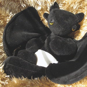 May include: Black plush bat with white accents. The bat has large, yellow eyes and is lying on a brown furry surface.