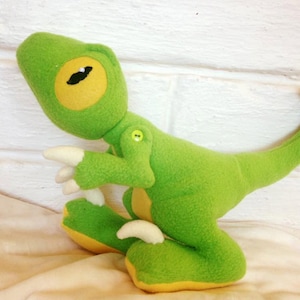 Puede incluir: Juguete de dinosaurio de peluche verde con un ojo amarillo y un botón en la espalda.