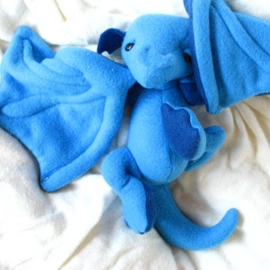 Op de afbeelding: Een blauw pluche draken speelgoed met vleugels en een staart. De draak ligt op een wit deken.