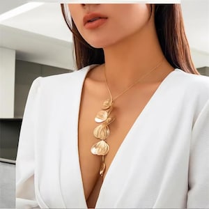 Peut inclure: Un collier doré avec un pendentif composé d'une série de formes stylisées en forme de feuilles. Le collier est porté sur un haut blanc, créant un contraste qui met en valeur le design du bijou. Le pendentif descend le long du décolleté.