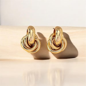 Peut inclure: Une paire de boucles d'oreilles dorées. Chaque boucle d'oreille présente un grand anneau arrondi en haut, relié à un ensemble de boucles métalliques torsadées et entrelacées. Les boucles d'oreilles sont présentées sur une surface en bois clair, avec une ombre douce.