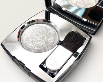 Könnte beinhalten: Ein quadratisches schwarzes Kompaktgehäuse mit Klappdeckel, das ein rundes Pfännchen mit schimmerndem weißem Puder-Highlighter und einen kleinen Applikatorpinsel mit dunklen Borsten enthält. Das Innere hat einen silbernen Rahmen und einen Spiegel.
