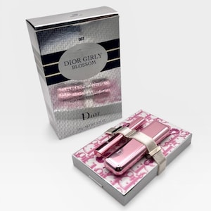 May include: A silver box and a pink makeup set. The box has the text "DIOR GIRLY BLOSSOM" and "READY-TO-WEAR SUMMER MAKEUP". The makeup set includes a pink lipstick, a pink applicator, and a pink compact, all held together with a silver band.