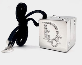 May include: A silver-toned metallic cube charm featuring the letters Dior encrusted with clear crystals on the front face. The cube is attached to a black fabric cord with a silver metal clasp at the end, designed for hanging or attachment.