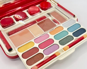 May include: A red makeup kit with a variety of cosmetics. The kit includes eyeshadows in various colors, lipsticks, blush, and applicators. The kit is open, revealing the different makeup products inside.