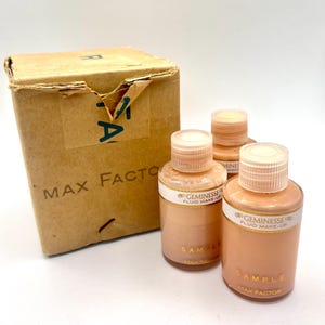 May include: A vintage Max Factor box with the text "MAX FACTOR" and three bottles of Geminesse Fluid Make-Up. The bottles are labeled "SAMPLE" and have a light beige liquid inside. The box is made of cardboard.