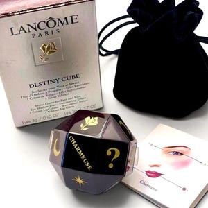 May include: A Lancôme Destiny Cube makeup set featuring a metallic box with gold rose logo, a dark purple velvet drawstring pouch, and a multi-faceted cube container with gold symbols including a crescent moon, star, and question mark.
