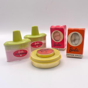 May include: A collection of vintage Barbie miniature vanity accessories including two clear plastic face powder containers with lime green lids, a round yellow rouge tin, a pink box labeled Powder Puffs, and an orange box labeled Cotton Balls.