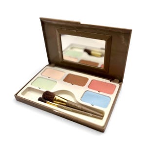 May include: Open brown makeup palette with a mirror in the lid. The palette contains six different colored makeup products, including pink, green, brown, and blue. Two makeup brushes are included in the palette.