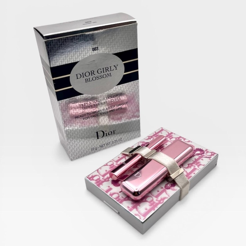 May include: A silver box and a pink makeup set. The box has the text "DIOR GIRLY BLOSSOM" and "READY-TO-WEAR SUMMER MAKEUP". The makeup set includes a pink lipstick, a pink applicator, and a pink compact, all held together with a silver band.