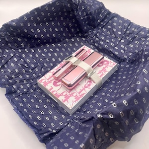 May include: A pink and silver makeup set, including a compact, two applicators, and a small notebook, is presented on a patterned box. The set is secured with a silver ribbon and rests on blue tissue paper with a repeating pattern.