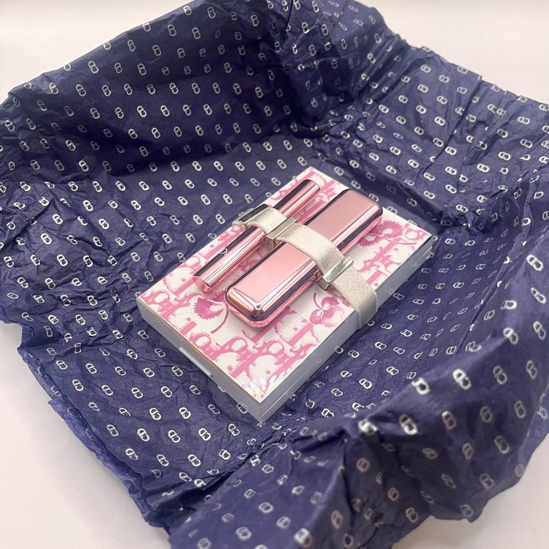 May include: A pink and silver makeup set, including a compact, two applicators, and a small notebook, is presented on a patterned box. The set is secured with a silver ribbon and rests on blue tissue paper with a repeating pattern.